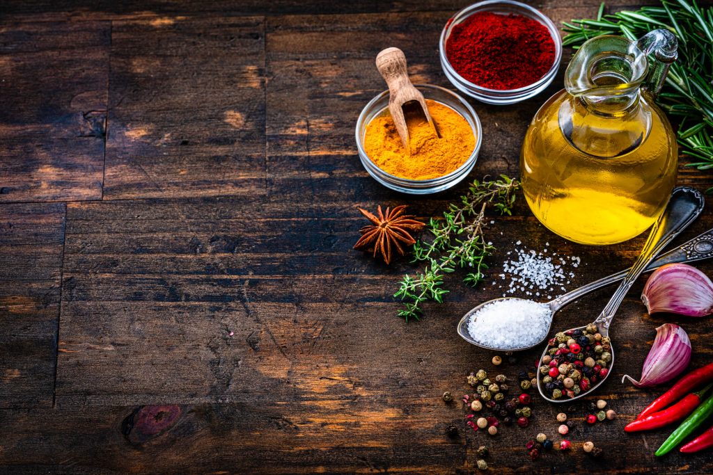 high angle view of a bottle with extra virgin olive oil, two spoons with peppercorns and salt, two bowls with paprika and turmeric arranged at the right of a rustic wooden table