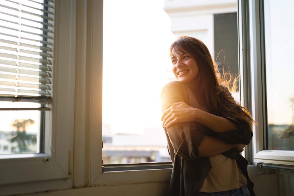 Beautiful young woman looking through the window while enjoying fresh air and sunlight